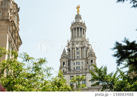 Tall historic tower featuring a golden statue on top rises above green trees in a downtown city center. The detailed architecture stands prominently among modern urban buildings. Tall historic tower featuring a golden statue on top rises above green trees in a downtown city center. The detailed architecture stands prominently among modern urban buildings. 138083488