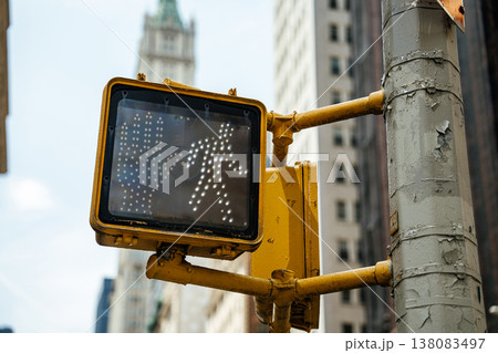 Close up of a pedestrian walk signal mounted on a traffic pole in a busy urban city with tall skyscrapers in the background. The illuminated walking figure indicates safe street crossing in a downtown Close up of a pedestrian walk signal mounted on a traffic pole in a busy urban city with tall skyscrapers in the background. The illuminated walking figure indicates safe street crossing in a downtown 138083497