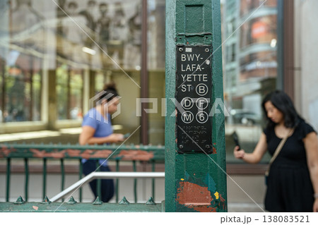 A Broadway Lafayette Street subway sign displays multiple train lines at a New York City entrance. Pedestrians using smartphones pass by showing modern city commuting behavior. A Broadway Lafayette Street subway sign displays multiple train lines at a New York City entrance. Pedestrians using smartphones pass by showing modern city commuting behavior. 138083521