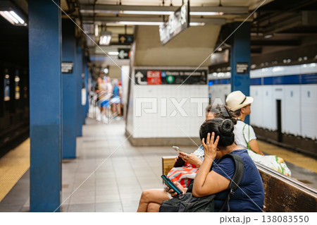 Woman using smartphone while sitting on bench at Broadway Lafayette Street subway station in New York City. Urban underground metro platform scene showing daily commute, tiled walls, and public 138083550