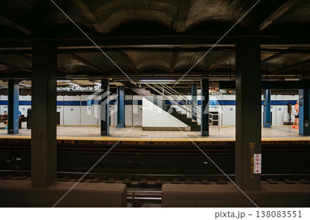 Symmetrical underground view of Broadway Lafayette Street subway station platform in New York City. Urban metro scene with staircase, tiled walls, railway tracks, and industrial steel columns. 138083551
