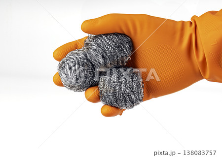 Hand wearing orange protective rubber glove holding three shiny stainless steel wool scrubber pads for heavy-duty kitchen cleaning and dishwashing tasks on white background 138083757