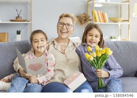 Smiling mature woman sitting on sofa, hugging kids congratulating her with tulips and handmade card. Smiling mature woman sitting on sofa, hugging kids congratulating her with tulips and handmade card. 138084546