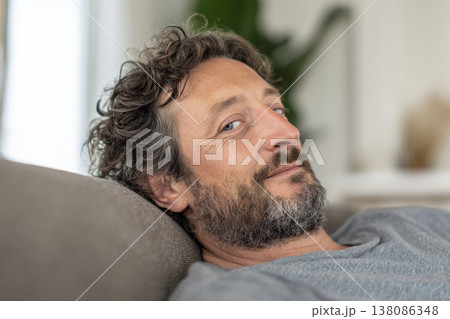 man resting on sofa with gentle expression, bright contemporary living room, soft natural sunlight. 138086348