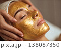 close-up of female client with radiant skin covered in gold face mask, esthetician gently adjusting 138087350