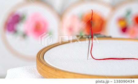 A thin silver needle is moving through bright crimson silk thread and white fabric, showing a detail of the embroidery process. Background features colorful floral designs. 138090434