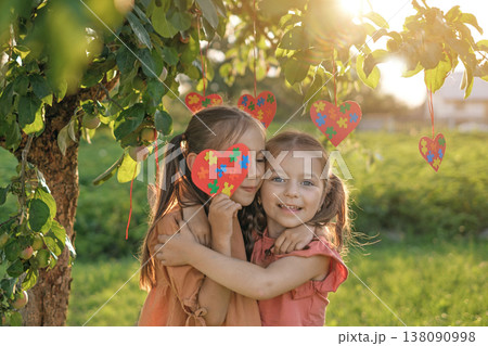 Two little girls hugging under apple tree holding colorful heart with puzzle pattern at sunset. Concept of autism awareness, friendship, childhood love and support. Two little girls hugging under apple tree holding colorful heart with puzzle pattern at sunset. Concept of autism awareness, friendship, childhood love and support. 138090998