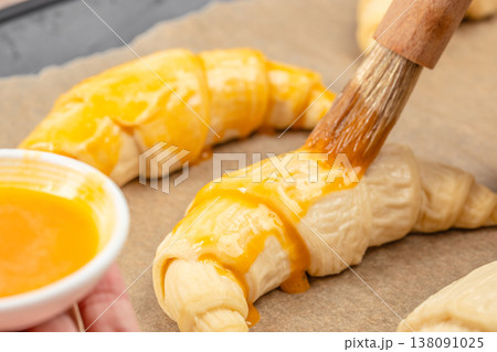 Raw croissant brushed with egg wash on baking paper, close-up pastry preparation process Raw croissant brushed with egg wash on baking paper, close-up pastry preparation process 138091025