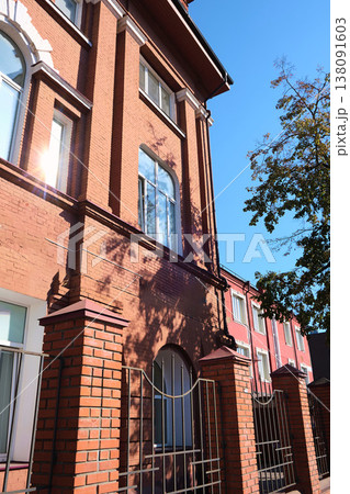 Facade of an old red brick building with arched windows and decorative white accents, partially shaded by trees and sunlight reflections. The image is suitable for topics about building restoration Facade of an old red brick building with arched windows and decorative white accents, partially shaded by trees and sunlight reflections. The image is suitable for topics about building restoration 138091603