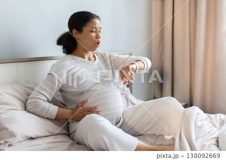 Pregnant Black woman sitting on bed timing contractions, checking watch 138092669