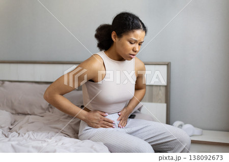 Young black woman sitting on bed holding lower abdomen in pain Young black woman sitting on bed holding lower abdomen in pain 138092673