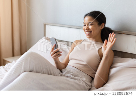 Young black woman lying on bed waving at smartphone during video call 138092674