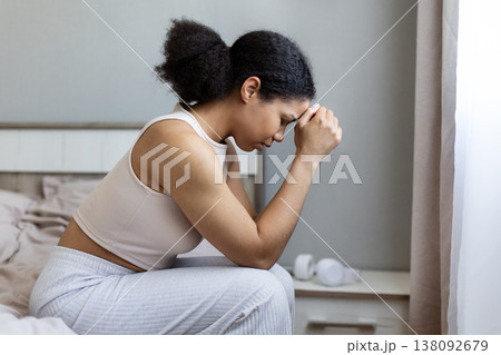 Upset black woman sitting on bed with hands clasped and head down Upset black woman sitting on bed with hands clasped and head down 138092679