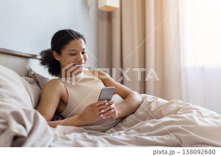 Young black woman lying in bed smiling while using smartphone 138092680