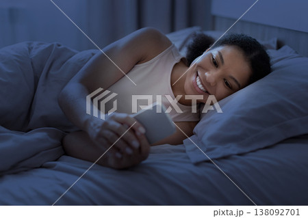 Young black woman lying in bed at night smiling while using smartphone 138092701