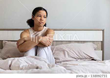 Black woman sitting on bed hugging knees, looking away with sad expression Black woman sitting on bed hugging knees, looking away with sad expression 138092731