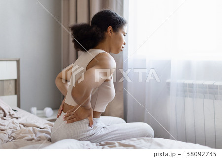Young african american woman sitting on bed holding lower back in pain Young african american woman sitting on bed holding lower back in pain 138092735