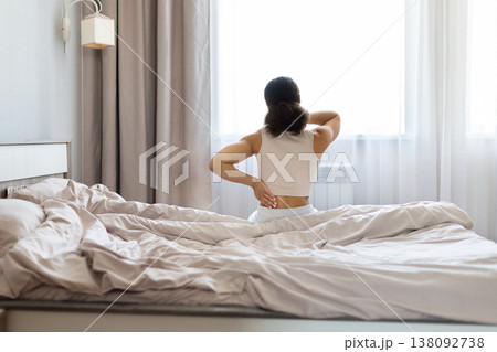 Black woman sitting on bed holding neck and lower back in discomfort Black woman sitting on bed holding neck and lower back in discomfort 138092738