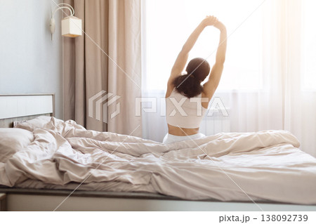 Young black woman stretching arms overhead while sitting on bed, enjoying morning Young black woman stretching arms overhead while sitting on bed, enjoying morning 138092739