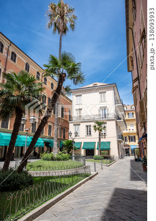 Alassio's charming town square features lush palm trees, vibrant green awnings on buildings, and traditional italian architecture under a bright blue sky on a sunny summer day 138093933