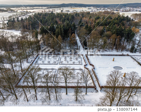 Aerial view of a symmetrical formal garden covered in snow. A majestic winter landscape featuring geometric hedges, frozen fountains, and a dense pine forest under a cloudy seasonal sky. Aerial view of a symmetrical formal garden covered in snow. A majestic winter landscape featuring geometric hedges, frozen fountains, and a dense pine forest under a cloudy seasonal sky. 138097144
