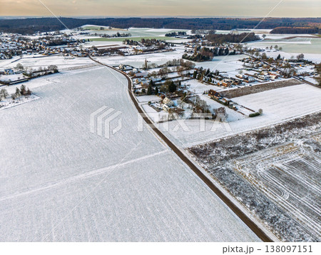 Aerial view of a narrow road winding through snow covered fields and a rural European village. A minimalist winter landscape with frosted farmland, scattered houses, and a distant forest line. 138097151