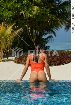 A beautiful tanned girl with an athletic physique in a swimsuit looks at the sea through palm trees on the edge of the villa pool on a sunny day in the Maldives, she is happy and posing A beautiful tanned girl with an athletic physique in a swimsuit looks at the sea through palm trees on the edge of the villa pool on a sunny day in the Maldives, she is happy and posing 138097655