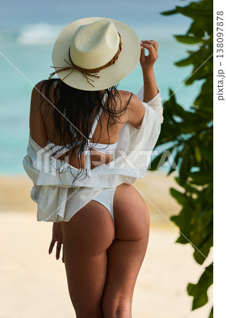 A beautiful tanned girl is wearing a hat and swimsuit with an athletic physique stands on the beach of the villa on a sunny day in the Maldives, she is happy and posing 138097878