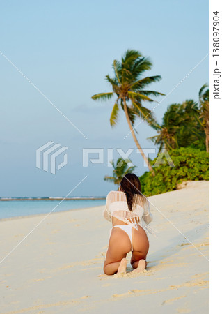 A beautiful tanned girl in a swimsuit sits along a white beach at sunset in the Maldives, she is happy, posing for a photographer 138097904