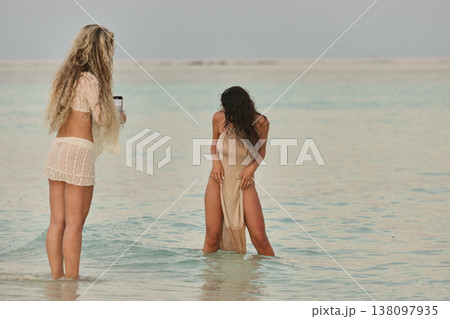Two beautiful tanned girl friends in a swimsuit taking pictures of each other, along a white beach at sunset in the Maldives, she is happy, she is posing 138097935