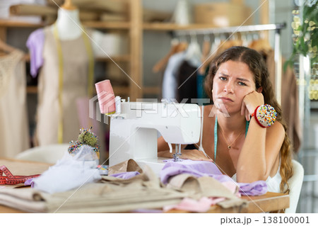 Sad dressmaker sitting at sewing machine in studio 138100001