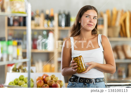 Young female shopper carefully choosing jar of canned olives in grocery supermarket 138100113