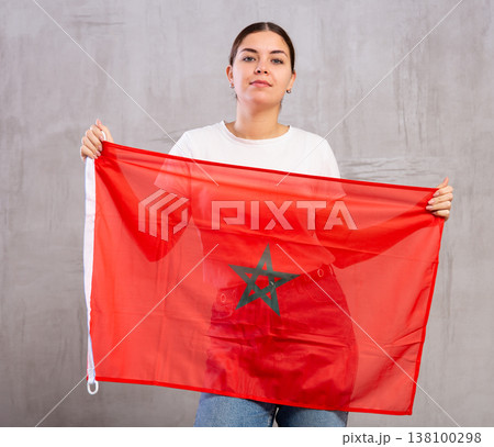 Calm cheerleader girl holds Moroccо flag in her hands. Isolated on gray background Calm cheerleader girl holds Moroccо flag in her hands. Isolated on gray background 138100298