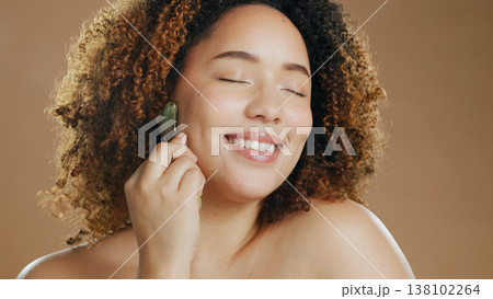 Happy woman, relax or jade roller in studio for face with anti aging product or facial tool on brown background. Smile, beauty or confident girl model with cosmetics for wellness or natural skincare Happy woman, relax or jade roller in studio for face with anti aging product or facial tool on brown background. Smile, beauty or confident girl model with cosmetics for wellness or natural skincare 138102264