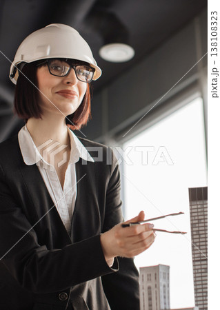 A female architect wearing a hard hat and glasses holds dividers near a building model, symbolizing construction and design planning 138108323