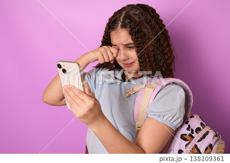 Emotional Young Girl with Curly Hair Crying While Holding a Smartphone Against Purple Background Emotional Young Girl with Curly Hair Crying While Holding a Smartphone Against Purple Background 138109361