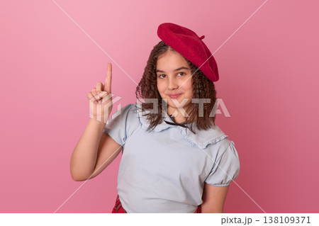 A Young Girl in a Beret Playfully Posing Against a Pink Background with a Finger Raised 138109371