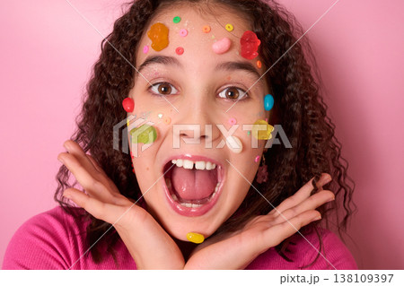 Joyful Expression of a Young Girl Surrounded by Colorful Candy on Her Face Against Pink Background Joyful Expression of a Young Girl Surrounded by Colorful Candy on Her Face Against Pink Background 138109397