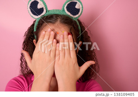 Child Hiding Face with Hands Wearing Fun Frog Headband Against a Soft Pink Background Child Hiding Face with Hands Wearing Fun Frog Headband Against a Soft Pink Background 138109405