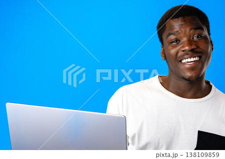 Man works on laptop and smiles in front of bright blue background during daytime Man works on laptop and smiles in front of bright blue background during daytime 138109859