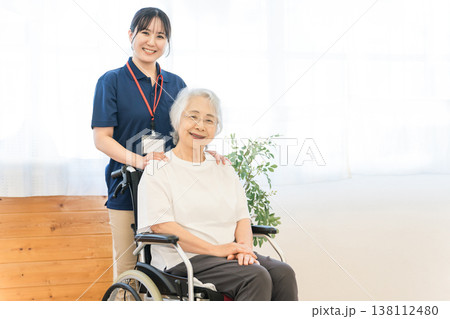 車椅子に乗るシニア女性と介護施設で働く女性スタッフ 車椅子に乗るシニア女性と介護施設で働く女性スタッフ 138112480