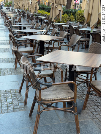 Empty outdoor cafe terrace with aligned wicker chairs and tables under folded umbrellas. Off peak dining setup, hospitality readiness, street seating arrangement, business downtime, urban service 138114337