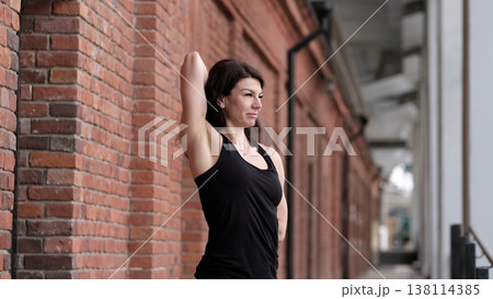 A woman performs a standing stretch, arms behind her back, leaning on a brick wall. This scene highlights the dedication to flexibility exercises and the importance of taking time for self-care. 138114385