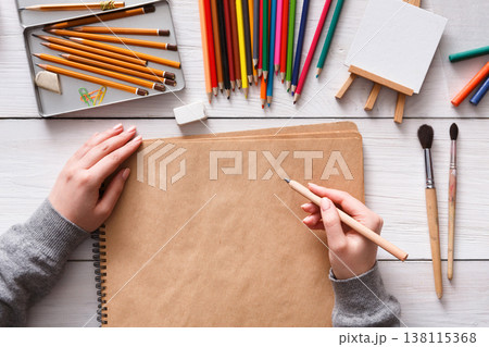 Top view of artist's workplace - hands with pencil on wooden desktop with blank paper and drawing tools Top view of artist's workplace - hands with pencil on wooden desktop with blank paper and drawing tools 138115368