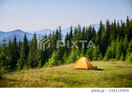 Bright orange tourist tent on grassy hillside, surrounded by lush green pine trees. Breathtaking view of distant blue mountains under clear sky, creating idyllic and serene camping spot. Bright orange tourist tent on grassy hillside, surrounded by lush green pine trees. Breathtaking view of distant blue mountains under clear sky, creating idyllic and serene camping spot. 138115959