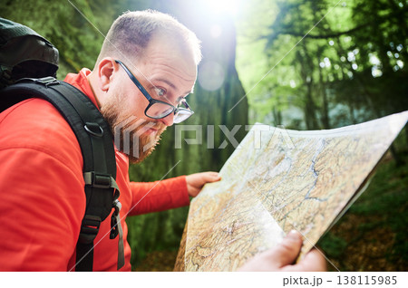 Close up of bearded tourist man studies map while stands in front of large rock in dense forest. Traveler with grey backpack and glasses, planning outdoor adventure, hiking or climbing route. Close up of bearded tourist man studies map while stands in front of large rock in dense forest. Traveler with grey backpack and glasses, planning outdoor adventure, hiking or climbing route. 138115985