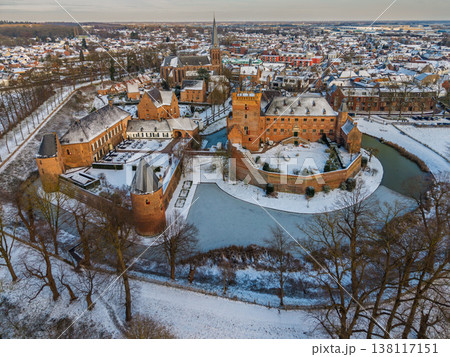 Aerial view of a medieval brick castle with a water filled moat in a snowy European town. Picturesque winter landscape featuring historical architecture, a church spire, and frosted rooftops. Aerial view of a medieval brick castle with a water filled moat in a snowy European town. Picturesque winter landscape featuring historical architecture, a church spire, and frosted rooftops. 138117151