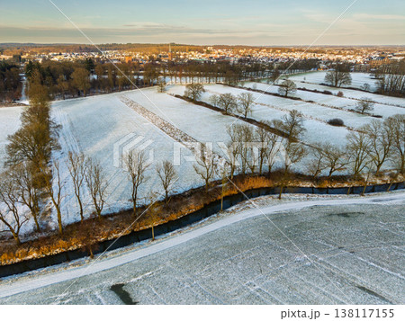 Aerial view of a snowy Dutch landscape at sunset. A small canal winds through frost-covered fields and rows of bare trees, with a charming European village and church spire on the golden horizon. Aerial view of a snowy Dutch landscape at sunset. A small canal winds through frost-covered fields and rows of bare trees, with a charming European village and church spire on the golden horizon. 138117155