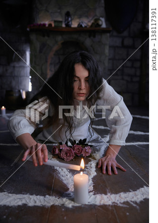 Woman with dark hair meticulously outlines sacred symbol on floor surrounded by candles and flowers Woman with dark hair meticulously outlines sacred symbol on floor surrounded by candles and flowers 138117391