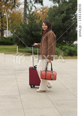A stylish woman prepares to travel, holding luggage near a modern building. The image combines fashion, confidence, and the mood of departure. A stylish woman prepares to travel, holding luggage near a modern building. The image combines fashion, confidence, and the mood of departure. 138119030
